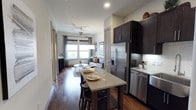 This thoughtfully designed single-floor space showcases an open-concept layout that seamlessly connects the kitchen, dining, and living areas into one cohesive and functional environment. Rich, dark hardwood flooring flows throughout, anchoring the space with warmth and sophistication.

The kitchen is appointed with dark espresso cabinetry that extends to the upper walls, stainless steel appliances including a French door refrigerator and dishwasher, and a sleek light-toned countertop island that doubles as a casual dining surface with barstool seating. A tile backsplash adds texture and contrast to the kitchen's refined aesthetic.

Adjacent to the kitchen, the dining area centers around a modern table set for two, illuminated by a contemporary pendant light that draws the eye upward. The open living area beyond is bathed in natural light from large windows dressed with layered curtains, creating a bright and welcoming atmosphere. A ceiling fan with a light fixture provides both comfort and ambient illumination.

The living room features a comfortable sofa accented with decorative pillows, anchored by a patterned area rug. Framed artwork adorns the walls, adding a curated, homely touch. The overall ambiance is modern and inviting, blending dark and light tones for a balanced, contemporary aesthetic.