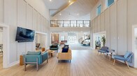 Located at 4705 Windsong Dr in Virginia Beach, VA, this impressive two-story amenity space spans a generous total floor area across two well-appointed levels. The main floor commands immediate attention with a soaring double-height lobby, flooded with natural light through expansive windows and anchored by a striking open-tread staircase with sleek black metal railings. The ground level hosts multiple distinct zones, including a spacious lounge area with stylish seating arrangements, a decorative latticed accent wall, and a well-equipped kitchenette bar featuring a white island with teal barstools, tile backsplash, and a mounted TV. A dedicated game area with a pool table and shuffleboard adds a recreational dimension, while a private office nook with a built-in workstation provides a quiet workspace. The upper mezzanine level offers an airy open layout overlooking the lobby below, complete with casual seating, woven pendant lighting, a foosball table, and a secondary lounge area. The hallway connecting spaces features framed artwork and additional soft seating. Throughout, warm wood-toned ceiling accents, recessed lighting, and a neutral palette of soft grays, blush, and tan create a modern, welcoming ambiance.