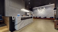 This two-floor commercial space is defined by a refined industrial aesthetic that blends upscale materials with contemporary design throughout. The ground floor welcomes visitors with a commanding reception desk clad in white Calacatta marble, accented by elegant brass-framed pendant lighting overhead. A feature wall of geometric patterned white tile serves as a dramatic backdrop to a long timber bench seat adorned with navy cushions, creating a formal yet inviting entry area with polished concrete floors underfoot.

Adjacent lounge areas on the ground floor offer varied seating arrangements, including a cognac leather sofa paired with a sleek black accent chair against a richly textured dark ceramic tile wall, and a large navy sectional sofa set beneath a warm floor lamp. Tufted leather banquette booths with round timber tables are tucked beneath the mezzanine overhang, illuminated by oversized dome pendants that create an intimate atmosphere.

The second floor, accessed via a black steel staircase, features an open mezzanine with black metal balustrades overlooking the floor below. This level offers additional lounge and dining seating alongside frosted glass-partitioned office areas. Dedicated meeting rooms are equipped with whiteboards, wall-mounted displays, natural timber tables, and bright accent chairs, providing functional spaces for collaboration throughout.