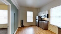 This single-floor space combines classic architectural character with modern updates to create a functional and inviting living environment. The kitchen is a standout feature, equipped with stainless steel appliances including a full-size refrigerator, gas range, and over-the-range microwave. Warm-toned, two-tone cabinetry pairs upper white cabinets with lower wood-finish units, offering ample storage. The dark countertops provide a sleek contrast to the overall light and airy aesthetic. Warm hardwood-style flooring runs throughout, adding a natural, welcoming feel underfoot. A large window dressed with white blinds allows natural light to fill the space, creating a bright and open atmosphere. A classic cast-iron radiator beneath the window speaks to the building's historic character while providing efficient heating. The walls feature a subtle two-tone color scheme, with a muted sage green accent wall complementing the crisp white trim and crown molding detail that frames the ceiling. The combination of updated fixtures and timeless architectural details gives the space a balanced, well-maintained character suited for everyday living.