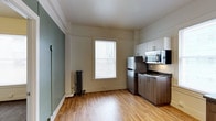 This single-floor space combines classic architectural character with modern updates to create a functional and inviting living environment. The kitchen is a standout feature, equipped with stainless steel appliances including a full-size refrigerator, gas range, and over-the-range microwave. Warm-toned, two-tone cabinetry pairs upper white cabinets with lower wood-finish units, offering ample storage. The dark countertops provide a sleek contrast to the overall light and airy aesthetic. Warm hardwood-style flooring runs throughout, adding a natural, welcoming feel underfoot. A large window dressed with white blinds allows natural light to fill the space, creating a bright and open atmosphere. A classic cast-iron radiator beneath the window speaks to the building's historic character while providing efficient heating. The walls feature a subtle two-tone color scheme, with a muted sage green accent wall complementing the crisp white trim and crown molding detail that frames the ceiling. The combination of updated fixtures and timeless architectural details gives the space a balanced, well-maintained character suited for everyday living.