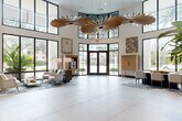 Located at 75 NE Sixth Ave in Delray Beach, Florida, this elegantly designed lobby space makes a striking first impression with its dramatic double-height ceilings and expansive floor plan. Large floor-to-ceiling windows and glass entry doors flood the interior with natural light, offering open views of the surrounding Florida landscape, including mature palm trees and lush greenery.

The architectural centerpiece of the space is a set of sculptural, sunburst-style wooden chandeliers suspended from the soaring ceiling, adding a bold yet refined artistic statement. Large-format light stone tile flooring extends throughout, providing a clean, polished foundation that enhances the sense of openness.

The lobby is thoughtfully zoned into distinct functional areas. A welcoming seating area near the entrance features contemporary neutral-toned furniture arranged on patterned area rugs. A more intimate lounge zone includes a generous sectional sofa, organic-shaped coffee tables, and floor-to-ceiling botanical-print drapery that frames sweeping outdoor views. Lush tropical planters serve as natural dividers, adding vibrant greenery to the interior.

Additional amenities visible within the space include a built-in media wall with custom cabinetry, a glassed-in private room, open shelving with curated décor, and an upper-level mezzanine with glass railings. The overall ambiance is modern, airy, and resort-inspired, consistent with the coastal character of Delray Beach.