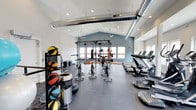 This single-floor fitness center is thoughtfully designed to accommodate a wide range of workout styles and training needs. The space is anchored by a striking vaulted ceiling with a soft blue accent wall, lending an open, airy atmosphere that enhances the overall workout experience. Exposed industrial-style ductwork runs along the ceiling, adding a modern edge to the clean, white interior.

Natural light pours in through multiple windows positioned along the walls, creating a bright and energizing environment. Recessed lighting supplements the natural light, ensuring the space remains well-lit throughout.

The layout is efficiently organized into distinct training zones. A row of treadmills and cardio machines lines one side, offering members a dedicated area for endurance training. At the center and rear of the space, a heavy-duty squat and power rack provides a focal point for strength training. A ceiling-mounted punching bag adds variety for combat and cardio conditioning. Near the entrance, a well-stocked rack of medicine balls in various sizes supports functional and dynamic training routines.

The flooring throughout is durable rubber matting, providing traction, cushioning, and noise reduction appropriate for high-intensity use. Ceiling fans ensure proper air circulation, contributing to a comfortable training environment. Overall, the space is practical, modern, and versatile.