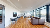 This contemporary single-floor amenity space is designed with an open-concept layout that encourages both relaxation and social interaction. Rich, warm-toned hardwood flooring extends throughout the entire area, providing a cohesive and inviting foundation. Crisp white walls and recessed ceiling lighting contribute to a clean, modern aesthetic, while abundant natural light pours in through expansive floor-to-ceiling windows that line one side of the space, offering views of an outdoor terrace and the surrounding environment beyond.

The main lounge area is furnished with a generously sized sectional sofa, accent chairs, and a low coffee table, creating a comfortable gathering zone. A large wall-mounted flat-screen television anchors one end of the room, accompanied by additional seating including woven accent chairs. A staircase with a sleek handrail connects to adjacent levels. Double exit doors with glazed panels provide access to additional areas. A dedicated kitchenette features a dark countertop, tile backsplash, built-in cabinetry with light wood finishes, a compact refrigerator, microwave, and a generous upper shelf — ideal for casual entertaining or day-to-day convenience. The overall ambiance is bright, airy, and thoughtfully designed for communal use.