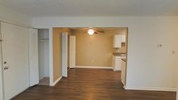 This single-floor residence offers a clean, functional layout designed for comfortable everyday living. The interior features rich, dark hardwood flooring that extends throughout the main living areas, providing a warm and grounded aesthetic that contrasts nicely with the crisp white trim and neutral warm-toned walls.

The open floor plan connects the living area seamlessly to the kitchen, creating an expansive, airy feel. The kitchen is outfitted with white cabinetry offering generous storage, dark stone-look countertops, and a sink positioned beneath upper cabinets — a practical and tidy arrangement. A ceiling fan with an integrated light fixture is centrally installed overhead, ensuring comfortable air circulation throughout the shared living and dining space.

Storage is a notable feature of this residence, with a dedicated closet area visible along one wall, complete with built-in shelving and hanging rods — ideal for organized storage needs. Bifold closet doors contribute to the clean, uncluttered look of the space.

The textured ceiling adds a subtle traditional touch, while the white baseboards and door frames provide crisp definition throughout. Overall, the space presents a straightforward, move-in-ready environment with a cohesive design that balances practicality with understated style.