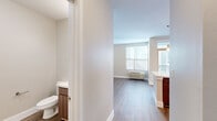This single-floor unit offers a clean, contemporary design highlighted by wood-look flooring that runs consistently throughout the space, complementing the neutral warm-beige and white walls. The kitchen is a standout feature, showcasing a large quartz-topped island with an undermount sink and modern pendant lighting above. Cabinetry in a warm wood-grain finish lines the walls, paired with a full suite of stainless steel appliances including a refrigerator, electric range, built-in microwave, and dishwasher. The layout transitions seamlessly from the kitchen into an open living area, which benefits from natural light streaming through horizontal-blind-covered windows. A sliding glass door provides direct access to an outdoor balcony, further enhancing the sense of openness. A wall-mounted HVAC unit serves the living space for individual climate control. The bathroom is efficiently designed, featuring a toilet and vanity with wood-toned cabinetry. An additional room off the main living area offers versatile use, also equipped with a window and wall HVAC unit. The overall ambiance is bright, modern, and move-in ready.