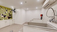 Located at 1777 Peachtree St NE in Atlanta, Georgia, this purpose-built pet spa facility is thoughtfully designed to provide a clean, comfortable, and fully equipped environment for pet grooming and bathing services. The space is finished with bright white walls and recessed lighting throughout, creating a hygienic and well-illuminated atmosphere.

The centerpiece of the room is a step-access bathing station, featuring built-in tiled stairs that allow pets to walk up to a sunken tub with ease. The surrounding walls in the bathing area are clad in classic white subway tile and a decorative lattice-patterned mosaic tile, adding both durability and visual interest. A wall-mounted hose and grooming dryer attachment are conveniently positioned near the tub for efficient use.

On the opposite side of the room, a built-in white cabinetry unit provides ample storage, topped with a countertop workspace for grooming tasks. A large wall-mounted industrial fan ensures proper air circulation, complementing the wall-mounted mini-split climate control system for year-round comfort. A vibrant floor-to-ceiling photographic mural depicting a variety of dogs on a grassy field adds a welcoming, playful touch to the space. A floor drain and carpeted areas in the prep zone complete the practical, pet-friendly design of this well-appointed grooming suite.