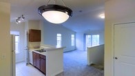 This single-floor space showcases an open-concept layout that seamlessly connects the kitchen and living areas, creating a sense of spaciousness and ease of movement. The kitchen is outfitted with dark wood cabinetry, a built-in dishwasher, and a double-basin sink set into a light-toned countertop. A peninsula-style counter extends into the space, offering a natural divide between the kitchen and the adjoining living area while maintaining visual openness. A prominent dome pendant light fixture hangs above the kitchen area, providing focused overhead illumination with a contemporary touch. The living area features soft wall-to-wall carpeting in a neutral tone, contributing to a comfortable and cohesive aesthetic. Recessed and flush-mount ceiling lights supplement the natural light that enters through multiple windows along the exterior walls. The windows, dressed with simple treatments, allow views of surrounding greenery and bathe the interior in natural light. The walls are finished in a clean, light neutral tone, enhancing the bright and airy atmosphere throughout. The overall design balances functionality with an inviting, comfortable ambiance suited for everyday living.