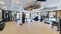This fitness center at 927–933 Main St in Sayreville, NJ, offers a thoughtfully designed workout space across a single open floor. The layout maximizes functionality, providing ample room for a wide variety of exercise equipment without feeling cramped. Large floor-to-ceiling windows line the perimeter, flooding the space with natural light and offering views of the surrounding outdoor environment, creating an energizing atmosphere for workouts.

The cardio zone features True-brand treadmills, stationary cycling bikes, a stair climber, and a rowing machine, all positioned to take advantage of the natural light from the expansive windows. A large wall-mounted flat-screen TV accompanies the cardio area for entertainment during workouts. The strength training section is equally well-appointed, housing a full rack of Hoist dumbbells, multiple Hoist cable and weight machines, adjustable benches, and a multi-functional cable cross system.

Floor-to-ceiling mirrors line one wall, visually expanding the space and allowing users to monitor their form. Ceiling fans with modern, dark-finished blades are distributed throughout, supplemented by recessed lighting and a distinctive geometric pendant light fixture that adds a contemporary design element. A neutral, textured carpet covers the floor throughout, providing a clean, professional finish. An AED unit is also present, reflecting a commitment to user safety.