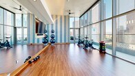 This contemporary fitness center is situated on an upper floor of a high-rise building, offering an inspiring workout environment defined by dramatic floor-to-ceiling glass walls that frame sweeping panoramic views of a dense urban skyline. The generous ceiling height and expansive glazing flood the space with natural light throughout the day, creating an open, energizing atmosphere.

The workout area features warm hardwood flooring throughout, providing a durable yet inviting surface suitable for a wide range of exercise activities. Accent walls clad in a textured blue-and-white striped wallcovering add a stylish, modern touch, while full-length mirrors along one wall enhance the sense of space and support proper form during workouts.

Equipment includes a lineup of stationary spin bikes positioned directly in front of the glass walls, offering riders unobstructed skyline vistas during sessions. A rack of kettlebells in varying sizes, a set of dumbbells, and stacked stability balls round out the free-weight and functional training options. A wall-mounted flat-screen television provides entertainment, and a ceiling fan ensures comfortable air circulation. Built-in shelving near the entry houses complimentary rolled towels for convenience. Glass double doors lead to an outdoor terrace, further connecting the fitness experience to the surrounding cityscape.