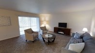This single-floor space offers a welcoming and well-appointed layout designed for comfortable everyday living. The living area serves as the heart of the space, featuring plush wall-to-wall carpeting that adds warmth underfoot. A large sliding glass door dressed with vertical blinds floods the room with natural light while providing a seamless connection to the outdoors. The living room is anchored by a sleek media console housing a flat-screen television and an integrated electric fireplace, offering both visual appeal and a cozy focal point. Neutral-toned walls complement the soft, understated color palette throughout, creating a calm and inviting ambiance. The seating arrangement — including a sofa, two accent armchairs, and a round coffee table — is thoughtfully arranged to encourage relaxed conversation and entertaining. A floor lamp provides additional ambient lighting, enhancing the room's warm atmosphere during evening hours. The overall design aesthetic is modern yet approachable, with clean lines and a cohesive neutral palette that lends the space a timeless, move-in-ready appeal. This space is well-suited for those seeking a straightforward, comfortable, and functional single-level living environment.