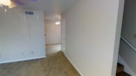 This two-floor property presents a straightforward, functional layout with neutral design throughout. The interior features wall-to-wall beige carpeting that extends across the rooms and connecting hallway, offering a warm and cohesive feel underfoot. Walls are finished in a clean off-white tone, complemented by white baseboards that provide a crisp contrast against the carpet.

A ceiling fan with a built-in light fixture is installed overhead, serving both ventilation and lighting needs. HVAC venting is integrated into the upper wall, indicating a central air system. The space benefits from a simple, open flow between areas, connected by a hallway with a white-paneled door featuring a brass-toned handle, adding a subtle traditional touch.

Electrical outlets are positioned at standard intervals along the walls, and the overall layout suggests a practical, livable design. The neutral palette and clean finishes throughout create a blank-canvas environment, making the space adaptable to a variety of furnishing styles. The two-floor configuration provides additional separation of living areas, enhancing the overall functionality of the property.