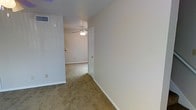 This two-floor property presents a straightforward, functional layout with neutral design throughout. The interior features wall-to-wall beige carpeting that extends across the rooms and connecting hallway, offering a warm and cohesive feel underfoot. Walls are finished in a clean off-white tone, complemented by white baseboards that provide a crisp contrast against the carpet.

A ceiling fan with a built-in light fixture is installed overhead, serving both ventilation and lighting needs. HVAC venting is integrated into the upper wall, indicating a central air system. The space benefits from a simple, open flow between areas, connected by a hallway with a white-paneled door featuring a brass-toned handle, adding a subtle traditional touch.

Electrical outlets are positioned at standard intervals along the walls, and the overall layout suggests a practical, livable design. The neutral palette and clean finishes throughout create a blank-canvas environment, making the space adaptable to a variety of furnishing styles. The two-floor configuration provides additional separation of living areas, enhancing the overall functionality of the property.