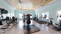 This fitness center offers a thoughtfully designed workout environment that balances functionality with an inviting aesthetic. The space is defined by its distinctive tray ceiling clad in warm natural wood planking, accented by recessed lighting and multiple ceiling fans that ensure comfortable air circulation throughout. Soft aqua-painted walls contribute a calm, refreshing atmosphere, while light-toned hardwood flooring provides a clean, durable surface underfoot.

The facility is well-stocked with a diverse range of equipment. A row of treadmills with individual monitor screens lines the windowed wall, allowing users to enjoy natural light during cardio sessions. An elliptical trainer complements the cardio area, while a multi-station cable and weight machine anchors the strength training zone. A tiered dumbbell rack stocked with various weights occupies a central position, alongside a weighted slam ball and protective floor mat for functional training. Additional resistance and conditioning equipment is visible along the perimeter.

Multiple windows on opposing walls allow ample natural light to fill the space, enhancing the open, airy feel. The overall design strikes a balance between a resort-inspired aesthetic and practical athletic functionality.