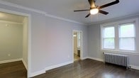 This single-floor space showcases a blend of classic architectural charm and functional design. The main room is finished with rich, dark hardwood floors that contrast cleanly against soft gray-painted walls and crisp white crown molding, which runs along the perimeter of the ceiling to add an elegant, traditional touch. A ceiling fan with a built-in light fixture is centrally mounted, providing both airflow and ambient lighting. A white cast-iron radiator positioned beneath a double-hung window with a view of greenery outside serves as a nod to the building's classic heritage while providing reliable heating. Natural light filters in through the window, brightening the space. A doorway leads to an adjoining room, visible through a white-trimmed opening, which also features a radiator and appears to receive natural light from its own window. White baseboards throughout complement the crown molding, reinforcing the cohesive, period-influenced aesthetic. The overall ambiance is clean, bright, and understated, with architectural details that speak to the character of a well-maintained, traditionally styled property.