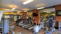 This fitness center is thoughtfully designed to provide a fully functional workout environment within a single-level layout. The space features a dedicated cardio zone lined with multiple treadmills and at least one elliptical machine, all from True Fitness — a reputable commercial-grade equipment brand. The machines are equipped with individual digital display consoles, allowing users to track their workout metrics with ease.

The interior design incorporates warm earth tones, highlighted by deep orange accent walls that add energy and visual interest to the space. Large wall-to-wall mirrors run along multiple walls, enhancing the sense of openness and allowing users to monitor their form during exercise. The ceiling is fitted with recessed and flush-mounted fluorescent lighting, ensuring the space is well-lit throughout.

A wall-mounted flat-screen television is positioned prominently for visibility from the cardio equipment, supporting an engaging workout experience. The flooring features a multi-toned patterned carpet that complements the warm color palette of the room. Additional exercise equipment is visible in the background, suggesting a broader range of workout options beyond the primary cardio area. Overall, the fitness center is functional, organized, and designed with the active user in mind.