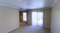 This single-floor space presents a clean and functional living area characterized by neutral, warm-toned walls and wall-to-wall carpeting that adds comfort underfoot. The room benefits from ample natural light, provided by a standard window on one wall and a set of full-length sliding glass doors fitted with vertical blinds, offering direct access to an outdoor area with visible greenery beyond. Crown molding frames the textured popcorn ceiling, adding a subtle architectural detail that elevates the overall finish of the space. A recessed ceiling light provides additional overhead illumination. The open layout allows for flexible furniture arrangements, making it well-suited for use as a living or dining area. Along one wall, built-in open shelving offers convenient storage or display space. White baseboards run throughout, providing a crisp contrast to the neutral carpet and wall tones. The overall ambiance is understated and practical, with a straightforward design that prioritizes functionality and natural light.