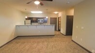 This single-floor space showcases an open-concept layout that seamlessly connects the kitchen and living area. The kitchen is appointed with dark espresso-toned cabinetry that extends to the upper walls, providing generous storage. A wide peninsula with light granite-style countertops and bar-height seating divides the kitchen from the living space, making it ideal for casual dining or entertaining. Stainless steel appliances, including a refrigerator and microwave, are integrated into the design. Pendant lighting hangs above the peninsula, complementing the ceiling fan with light fixture that anchors the living area. The flooring transitions from hard surface in the kitchen to plush neutral carpet in the living space. Warm beige walls create a welcoming ambiance throughout. A hallway visible in the background leads to additional rooms, with what appears to be a walk-in closet and additional doorways offering further functionality. The overall layout is practical and well-organized, with consistent dark-toned door and trim details tying the design together cohesively.