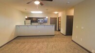 This single-floor space showcases an open-concept layout that seamlessly connects the kitchen and living area. The kitchen is appointed with dark espresso-toned cabinetry that extends to the upper walls, providing generous storage. A wide peninsula with light granite-style countertops and bar-height seating divides the kitchen from the living space, making it ideal for casual dining or entertaining. Stainless steel appliances, including a refrigerator and microwave, are integrated into the design. Pendant lighting hangs above the peninsula, complementing the ceiling fan with light fixture that anchors the living area. The flooring transitions from hard surface in the kitchen to plush neutral carpet in the living space. Warm beige walls create a welcoming ambiance throughout. A hallway visible in the background leads to additional rooms, with what appears to be a walk-in closet and additional doorways offering further functionality. The overall layout is practical and well-organized, with consistent dark-toned door and trim details tying the design together cohesively.