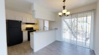This single-floor residence showcases an open-concept layout that connects the kitchen and dining area in a bright, cohesive living space. The kitchen is outfitted with white cabinetry, granite-look countertops, and black appliances, including a range with an overhead exhaust hood. A peninsula with a built-in sink and matching countertop serves as both a functional workspace and a natural divider between the kitchen and dining area, enhancing the open flow of the space. The light grey wood-look flooring runs throughout, adding a clean, contemporary feel underfoot. The dining area is highlighted by an elegant chandelier with warm-toned glass shades, lending a classic touch to the otherwise modern aesthetic. Large sliding glass doors with vertical blinds allow abundant natural light to flood the space and provide direct access to an outdoor area, where mature trees are visible, adding a sense of greenery and tranquility. The white walls and high ceiling further enhance the airy, open atmosphere of the space, making it feel welcoming and spacious.