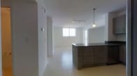 This single-floor residence showcases a clean, modern design centered around an open-concept floor plan that seamlessly connects the kitchen and living areas. The kitchen is anchored by a substantial dark-toned island with ample cabinetry in a rich wood-grain finish, providing both generous storage and a functional workspace. A stylish pendant light hangs above the island, complementing the track lighting system installed on the ceiling, which allows for flexible and adjustable illumination throughout the space. The flooring throughout is finished in large-format light ceramic tiles, contributing to a bright and cohesive aesthetic that extends across the entire layout. The living area is bathed in natural light, courtesy of generous windows that invite the outdoors in and create a warm, welcoming ambiance. The walls are finished in crisp white throughout, enhancing the sense of openness and making the space feel expansive. The overall design philosophy leans toward minimalism, with clean lines and a neutral palette that provides a versatile backdrop suited to a variety of interior styles. The layout flows intuitively, offering a comfortable and functional living environment ideal for everyday use.