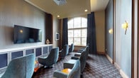 This single-floor communal lounge space is thoughtfully designed to offer both comfort and a polished aesthetic. The room's most striking architectural feature is a large arched window that floods the interior with natural light while framing views of a landscaped outdoor setting. Dramatic navy curtains frame the window, adding a sophisticated contrast to the warm neutral tones of the walls and ceiling.

Seating is arranged in a theater-inspired layout, with a collection of plush teal velvet swivel chairs positioned to face a wall-mounted flat-screen television. The TV is anchored above a long, low-profile credenza with matte navy cabinet doors and a clean white surface, accented by decorative ceramic vases on either side. Small orange accent tables are scattered among the seating, providing a vibrant pop of color throughout the space.

The walls are adorned with wood-framed textured panels and elegant crystal wall sconces that cast a warm, inviting glow. A patterned carpet in muted tones of grey and brown adds visual depth underfoot. Adjacent to the main seating area, glass-paneled doors lead to an adjoining space, reinforcing an open and connected feel throughout.