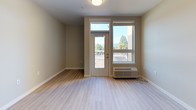 This thoughtfully designed single-floor unit offers a clean, contemporary aesthetic throughout. The open-concept kitchen and living area serves as the heart of the space, featuring light hardwood-style flooring that extends seamlessly through the main areas. The kitchen is well-appointed with warm wood-toned cabinetry, white countertops, and a full suite of stainless steel appliances including an electric range, dishwasher, and refrigerator, complemented by an under-cabinet range hood. A bright entry area provides convenient access from the front door, with additional cabinetry for storage nearby. The living area opens directly to a private balcony through a glass-paneled door, flanked by a large window that frames beautiful views of surrounding trees and mountains, filling the space with natural light. A light-filled hallway with durable hardwood-style flooring connects the main living area to the private rooms. The bedrooms feature soft carpeting, neutral warm-toned walls, and windows offering the same picturesque mountain and treetop scenery. The bathroom is finished with a light wood vanity, dark countertop, a large mirror, and a full soaking tub with shower, all set on clean tile flooring. The overall palette of warm neutrals and natural materials creates a calm, welcoming ambiance throughout.