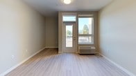 This thoughtfully designed single-floor unit offers a clean, contemporary aesthetic throughout. The open-concept kitchen and living area serves as the heart of the space, featuring light hardwood-style flooring that extends seamlessly through the main areas. The kitchen is well-appointed with warm wood-toned cabinetry, white countertops, and a full suite of stainless steel appliances including an electric range, dishwasher, and refrigerator, complemented by an under-cabinet range hood. A bright entry area provides convenient access from the front door, with additional cabinetry for storage nearby. The living area opens directly to a private balcony through a glass-paneled door, flanked by a large window that frames beautiful views of surrounding trees and mountains, filling the space with natural light. A light-filled hallway with durable hardwood-style flooring connects the main living area to the private rooms. The bedrooms feature soft carpeting, neutral warm-toned walls, and windows offering the same picturesque mountain and treetop scenery. The bathroom is finished with a light wood vanity, dark countertop, a large mirror, and a full soaking tub with shower, all set on clean tile flooring. The overall palette of warm neutrals and natural materials creates a calm, welcoming ambiance throughout.