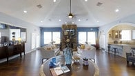 This inviting single-floor space makes a strong first impression with its soaring vaulted ceiling, which adds a sense of openness and grandeur to the main lounge area. A ceiling fan hangs at the apex, complementing the recessed lighting that illuminates the room evenly throughout. The focal point of the space is a striking floor-to-ceiling stacked stone fireplace, flanked by large windows dressed with white blinds that allow abundant natural light to pour in from multiple directions. Rich, dark hardwood flooring grounds the room and contrasts beautifully with the soft blue accent walls, creating a warm yet modern aesthetic. The lounge is thoughtfully arranged with plush, neutral-toned sofas and accent chairs accented with decorative throw pillows, fostering a comfortable and welcoming atmosphere. A large round central table serves as the heart of the seating area, styled with an ornate mosaic vase, floral arrangement, and reading materials. Decorative elements such as reed diffusers and ceramic accents add finishing touches throughout. The overall ambiance is bright, airy, and polished — a well-appointed communal space designed for both relaxation and casual interaction.