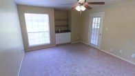 This single-floor space offers a welcoming and functional layout designed for comfortable living. The main room is generously proportioned, featuring soft wall-to-wall carpeting that adds warmth underfoot. A ceiling fan with an integrated light fixture is centrally mounted, providing both airflow and ambient lighting throughout the space.

Natural light is a defining characteristic of the room, with large windows fitted with horizontal blinds allowing for adjustable illumination. A glazed exterior door further enhances brightness while offering convenient access to the outdoors.

A built-in storage nook serves as a practical focal point along one wall, featuring open shelving above and white cabinetry with a dark countertop below. This compact built-in area provides useful storage and surface space, making the room both organized and versatile.

The walls are finished in a neutral beige tone, complementing the light carpeting and contributing to the room's calm, understated ambiance. White trim and baseboards frame the space cleanly, adding a crisp, finished look throughout. Overall, the space presents a straightforward, functional interior that is bright, open, and move-in ready.