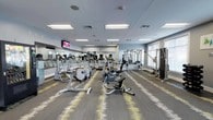 This fitness center offers a comprehensive workout environment designed to support a wide range of exercise routines. The main workout area is generously sized, featuring patterned carpet tiles in gray and yellow tones that add visual interest underfoot. Full-wall mirrors line one side of the room, enhancing the sense of space and allowing members to monitor their form during exercise. Large windows along the exterior walls invite natural light and provide outdoor views, creating an open and energizing atmosphere. The main floor is equipped with a robust selection of cardio machines, including upright and recumbent stationary bikes, ellipticals, and treadmills, along with multi-station cable machines and a dedicated free weights area featuring a dumbbell rack and kettlebells. Wall-mounted televisions are positioned throughout for entertainment during workouts. A vending machine provides convenient refreshment access. A separate studio or flex room features warm hardwood-style flooring, a wall-mounted TV, a large mirror, a medicine ball rack, and open floor space suitable for stretching, yoga, or group exercise. Motivational artwork decorates the walls throughout. The facility is illuminated by a combination of recessed and flush-panel ceiling lighting, ensuring a bright and functional environment throughout the day.