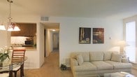 This single-floor residence offers a comfortable and cohesive layout designed for everyday living. The living area serves as the central gathering space, featuring plush wall-to-wall carpeting in a warm neutral tone that extends throughout the unit. A light beige sofa anchors the room, complemented by a pair of bold abstract wall art pieces that add a touch of color and personality to the otherwise understated décor. A low-profile baseboard heating unit is tucked neatly beneath the wall, and a ceiling vent ensures year-round climate control. Adjacent to the living area, a partial view into the kitchen reveals warm wood-toned cabinetry and a pass-through counter that maintains an open connection between the two spaces. A hallway extends from the living room, lined with smooth white walls and illuminated by a ceiling-mounted light fixture, leading to the remaining rooms at the rear of the unit. The overall ambiance is clean, calm, and functional, with a neutral color palette of whites and beiges creating a bright and cohesive feel throughout. The straightforward single-level layout ensures easy navigation and accessibility across all areas of the space.