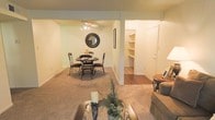 This single-floor residence offers a welcoming and functional layout designed for comfortable everyday living. The open-concept living and dining area is unified by warm, neutral-toned walls and soft wall-to-wall carpeting that creates a cozy atmosphere throughout. The dining area comfortably accommodates a round table and chairs, accented by a decorative round mirror and a ceiling fan with integrated lighting. Adjacent to the dining space, a transition to hardwood flooring leads to a built-in shelving alcove, offering excellent storage and display options. The built-in shelves are well-proportioned and add both practicality and architectural character to the space. A nearby door provides access to an additional room or closet, enhancing the home's overall storage capacity. The living area features a glass-top coffee table and plush seating, lending the space a relaxed and inviting feel. Subtle decorative touches, including potted greenery and candle accents, contribute to the warm ambiance. The overall design balances simplicity with comfort, making this a practical and livable home.