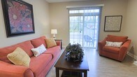This single-floor residence offers a comfortable and inviting layout designed for easy living. The living area serves as the heart of the space, bathed in natural light that streams through a large sliding glass door fitted with vertical blinds. The door opens to an outdoor patio, seamlessly connecting the interior to the outdoors and providing pleasant views of surrounding greenery. Wood-style flooring runs throughout, adding warmth and a sense of continuity to the space. The living room is thoughtfully arranged, with ample room for seating and a central coffee table, creating an ideal setting for both relaxing and entertaining. Neutral wall tones provide a versatile backdrop that complements the warm furnishings. Framed artwork accents the walls, contributing to the space's approachable and homey character. The overall ambiance is light, airy, and welcoming, with a practical, well-considered layout that makes efficient use of the available space. Natural light plays a key role in defining the atmosphere, keeping the interior feeling open and connected to the outdoors throughout the day.