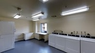 This single-floor laundry facility is designed with functionality and efficiency in mind. The room features a row of full-sized top-loading washing machines along one wall, each equipped with individual controls, providing ample capacity for multiple users. Complementing the machines is an L-shaped folding counter that runs along an adjacent wall, offering generous workspace for sorting and folding laundry. The flooring consists of durable grey vinyl, well-suited for a high-traffic utility space. Overhead, multiple fluorescent light fixtures ensure the room is brightly and evenly illuminated throughout. The textured ceiling adds a subtle architectural detail while contributing to the room's acoustics. A window on one wall allows for a degree of natural light and ventilation. The walls are finished in a neutral cream tone, keeping the space feeling clean and open. Overall, the facility presents a well-organized, practical environment equipped to serve the laundry needs of multiple occupants efficiently.