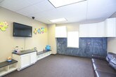 This single-story commercial space is thoughtfully designed to accommodate both children and adults, combining functional and engaging elements throughout. The main room serves as a children's activity area, featuring warm beige walls accented by colorful foam flower decorations and a wall-mounted flat-screen television. Built-in white cabinetry with a dark countertop provides ample storage, while a low bench with open cubby storage underneath offers seating and organization for toys and belongings. Plush stuffed animals and small toys add a welcoming, playful atmosphere.

A large chalkboard wall spans one side of the room beneath additional upper white cabinets, offering an interactive surface for children. The room is floored with neutral carpet throughout and illuminated by recessed drop-ceiling lighting panels.

A dark leather sofa faces a wide interior viewing window that looks directly into an adjacent fitness area equipped with exercise machines, allowing caregivers to supervise children while staying active. A solid wood door with a transom window above connects the space to a tiled entryway or corridor, completing the practical layout of this well-appointed facility.