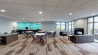 This well-appointed single-floor amenity space is thoughtfully divided into several distinct functional zones, offering a diverse range of activities and relaxation options. The open lounge area features a sleek linear fireplace set against a grey accent wall, flanked by floor-to-ceiling windows that flood the space with natural light and provide views of the surrounding outdoor greenery. A comfortable sectional sofa and side table complete this inviting retreat.

Adjacent to the lounge, a casual dining and coworking area is outfitted with round white tables and modern Eames-style chairs atop patterned wood-look flooring. A dedicated computer workstation nook, highlighted by a bold teal accent wall, houses multiple desktop computers and is well-suited for productivity tasks.

A glass-partitioned games room provides visual openness while defining a lively entertainment zone, complete with a pool table, ping-pong table, and shuffleboard. A separate gaming lounge features two wall-mounted flat-screen TVs — one connected to a PS4 — with seating positioned for an immersive experience. Throughout the space, recessed lighting, wood-look luxury vinyl flooring, drop ceilings, and wall-mounted air conditioning units contribute to a clean, contemporary aesthetic designed for both comfort and activity.