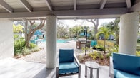 This inviting outdoor amenity space is thoughtfully designed to evoke a resort-like atmosphere, combining relaxation and recreation in a lush, tropical setting. At its centerpiece is a generously sized swimming pool with crystal-clear turquoise water, surrounded by a paved sun deck lined with lounge chairs and shaded by mature palm trees and native canopy trees that create a naturally cool environment.

A covered pavilion serves as the social hub of the space, featuring exposed beam ceilings, white textured columns, and smooth concrete flooring. The pavilion is furnished with a large sectional sofa in vibrant teal upholstery, complemented by side chairs and a coffee table — ideal for casual gatherings. A ceiling fan provides comfort during warmer months. Adjacent to the pavilion, a dedicated outdoor kitchen station equipped with a built-in grill offers convenient amenities for entertaining.

French doors lead from the covered patio directly into an interior clubhouse or leasing office, indicated by signage visible at the entry. On the opposite side of the pavilion, a tranquil pond with an active fountain provides a peaceful natural backdrop, framed by dense fern gardens and mature shade trees. The cohesive teal and neutral color palette throughout the furnishings and shade structures ties the entire outdoor space together with a polished, coastal aesthetic.