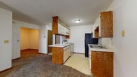 This single-floor residential unit offers a practical and functional layout designed for comfortable everyday living. The kitchen is positioned as a central hub of the home, featuring warm oak cabinetry paired with white upper cabinets, a white electric range, a black refrigerator, a dishwasher, and a double sink with light countertops. The kitchen flows seamlessly into the open living area, which is finished with plush wall-to-wall carpeting and neutral white walls that lend a clean, understated feel throughout. The spacious living area benefits from a baseboard heating system and provides generous room for a variety of furniture arrangements. A ceiling fan is also present in the space, adding to comfort and airflow. The bedroom is a quiet, carpeted room with ample wall space and straightforward finishes, offering a calm retreat. The full bathroom is well-appointed with a combined tub and shower unit, a toilet, and a single vanity sink set in a light-toned cabinet, complemented by tile flooring and chrome fixtures. A textured ceiling is consistent throughout the unit, adding subtle architectural character. The layout is efficient and open, making excellent use of the available space on a single level.