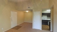 This single-floor space showcases an open-concept layout that connects the living area to the kitchen. The living area features neutral beige carpet and warm-toned walls, creating a clean and functional environment. A vaulted ceiling adds a sense of vertical space and airiness to the main living area, complemented by a ceiling fan fixture and a centrally placed overhead light. An HVAC vent is positioned along the ceiling, ensuring efficient climate control throughout the space.

A white interior door provides access to an additional room or closet off the main living area, while a second doorway leads toward a sunken section of carpeted flooring, adding a subtle architectural variation to the layout. Transitioning into the kitchen, the flooring shifts to hardwood-style planks, clearly delineating the two functional zones. The kitchen features dark-stained cabinetry, a mosaic tile backsplash in earthy tones, and a countertop workspace visible along the wall. The overall design maintains a straightforward, practical aesthetic with neutral finishes throughout.
