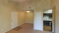 This single-floor space showcases an open-concept layout that connects the living area to the kitchen. The living area features neutral beige carpet and warm-toned walls, creating a clean and functional environment. A vaulted ceiling adds a sense of vertical space and airiness to the main living area, complemented by a ceiling fan fixture and a centrally placed overhead light. An HVAC vent is positioned along the ceiling, ensuring efficient climate control throughout the space.

A white interior door provides access to an additional room or closet off the main living area, while a second doorway leads toward a sunken section of carpeted flooring, adding a subtle architectural variation to the layout. Transitioning into the kitchen, the flooring shifts to hardwood-style planks, clearly delineating the two functional zones. The kitchen features dark-stained cabinetry, a mosaic tile backsplash in earthy tones, and a countertop workspace visible along the wall. The overall design maintains a straightforward, practical aesthetic with neutral finishes throughout.