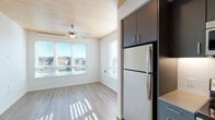 Located at 1090 NE 91st Ave in Hillsboro, Oregon, this thoughtfully designed single-floor unit showcases a contemporary aesthetic with warm, natural wood-paneled ceilings that serve as a defining architectural feature across the living spaces. Light-toned luxury vinyl plank flooring flows throughout the main living areas, complemented by crisp white walls that enhance the bright, airy atmosphere created by multiple windows with horizontal blinds allowing generous natural light.

The kitchen is a standout feature, offering two distinct configurations visible throughout the space. One version presents dark charcoal cabinetry paired with a stainless steel refrigerator and microwave, while another showcases deep navy-toned cabinetry with a full suite of stainless steel appliances including a range, dishwasher, and refrigerator. Both kitchens feature clean white subway tile backsplashes and ample upper and lower cabinet storage.

The bedroom areas include carpet flooring and ceiling fans, with windows overlooking outdoor greenery. The bathroom is well-appointed with a modern dark-toned vanity, quartz-style countertop, a large mirror with contemporary strip lighting, and direct access to a walk-in closet with built-in shelving and hanging rods. Recessed lighting and ceiling fans throughout add to the functional, modern character of this well-designed Hillsboro residence.
