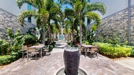 Located along NW Federal Highway in Stuart, Florida, this thoughtfully designed outdoor space serves as a central courtyard within a modern multi-story building complex. The architecture blends contemporary siding with natural stone veneer accents, creating a warm and visually appealing backdrop for the lush tropical landscaping that defines the space.

Towering royal palms and an array of tropical plantings—including banana plants, crotons, and manicured shrubs—frame the space with vibrant, year-round color. A central decorative stone fountain serves as a focal point, anchoring the paver courtyard and adding a tranquil water feature that enhances the ambiance. Symmetrically arranged outdoor dining tables and chairs flank the courtyard on both sides, offering inviting areas for gathering or relaxation beneath the open Florida sky.

Smooth concrete walkways wind through the grounds, lined with low-profile landscape lighting bollards that illuminate the pathways in the evening hours. Neatly mulched garden beds and gravel borders provide texture and contrast against the lush greenery. A manicured lawn area at the perimeter adds a soft, open green space to the overall composition.

The courtyard receives abundant natural sunlight and is framed on multiple sides by building balconies, creating an enclosed yet open-air retreat with a resort-like character throughout.