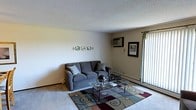 This single-floor space centers around a living room that offers a functional and comfortable layout. The room features a textured popcorn ceiling and neutral-toned walls that create a clean, understated backdrop. A wall-mounted air conditioning unit provides climate control, complemented by baseboard heating along the perimeter — practical features for year-round comfort. A large window dressed with vertical blinds runs along one wall, allowing a generous amount of natural light to fill the room while offering privacy options. The flooring transitions between carpet and hard surface near the entryway, adding definition to the space. Decorative wall art and a framed picture contribute a personalized touch to the otherwise simple wall surfaces. The living area comfortably accommodates a full-sized sofa with accent pillows, a glass-top coffee table with a metal frame, and a patterned area rug in earth tones, anchoring the seating arrangement. Overall, the space presents a straightforward, practical living environment with essential amenities in place.