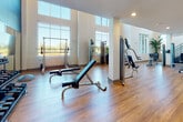 This two-story fitness center is designed to support a comprehensive workout experience, with equipment and dedicated zones spread across both floors. The main level is characterized by soaring ceilings and an abundance of natural light streaming through large, floor-to-ceiling windows that line the perimeter walls. Warm hardwood-style flooring runs throughout this level, lending a clean, modern aesthetic. Strength training equipment, including cable machines, benches, and a functional trainer, is arranged throughout the open floor plan. Rowing machines and additional resistance equipment occupy the central area, while a mirrored free weight zone with dumbbell racks and a flat-screen television creates a defined training section accentuated by a bold teal accent wall. A metal-railed staircase, flanked by a large tropical plant, connects to the upper level. The second floor houses a cardio suite equipped with treadmills, ellipticals, and cycling bikes, illuminated by contemporary linear pendant lighting and framed by windows offering outdoor views. A flexible exercise room with checkered tile flooring provides additional open training space, accompanied by restroom facilities and a small refreshment station nearby.