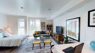 This single-floor unit offers a well-designed open-concept layout that integrates the living, sleeping, and dining areas into one cohesive space. Large floor-to-ceiling windows flood the interior with natural light while providing views of lush outdoor greenery, creating a bright and airy atmosphere throughout. The clean white walls and smooth ceilings complement the warm-toned hardwood flooring that runs consistently across the space.

The living area is thoughtfully arranged with mid-century modern furnishings, including a gray loveseat, a glass-top coffee table, and a wooden media console supporting a wall-mounted flat-screen television. Colorful artwork and a compact bookshelf add personality to the space. The sleeping area is situated adjacent to the living zone, separated by a partial wall that also provides closet storage with dual doors.

The kitchen is efficiently designed, featuring gray flat-front cabinetry, white subway tile backsplash, white quartz-style countertops, and a full suite of stainless steel appliances including a refrigerator, electric range, built-in microwave, and dishwasher.

The bathroom includes a combination tub and shower, a toilet, and a vanity with gray cabinetry and a wide mirror, maintaining the unit's consistent modern aesthetic with wood-look flooring.