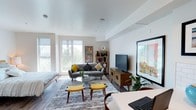 This single-floor unit offers a well-designed open-concept layout that integrates the living, sleeping, and dining areas into one cohesive space. Large floor-to-ceiling windows flood the interior with natural light while providing views of lush outdoor greenery, creating a bright and airy atmosphere throughout. The clean white walls and smooth ceilings complement the warm-toned hardwood flooring that runs consistently across the space.

The living area is thoughtfully arranged with mid-century modern furnishings, including a gray loveseat, a glass-top coffee table, and a wooden media console supporting a wall-mounted flat-screen television. Colorful artwork and a compact bookshelf add personality to the space. The sleeping area is situated adjacent to the living zone, separated by a partial wall that also provides closet storage with dual doors.

The kitchen is efficiently designed, featuring gray flat-front cabinetry, white subway tile backsplash, white quartz-style countertops, and a full suite of stainless steel appliances including a refrigerator, electric range, built-in microwave, and dishwasher.

The bathroom includes a combination tub and shower, a toilet, and a vanity with gray cabinetry and a wide mirror, maintaining the unit's consistent modern aesthetic with wood-look flooring.