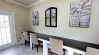 This single-floor space exudes a refined and welcoming ambiance, combining classic design elements with tasteful décor. The interior is finished with neutral, warm-toned walls complemented by crisp crown molding, lending a polished and cohesive look throughout.

A standout feature is the long, dark granite-topped dining or bar-height surface, elegantly supported by white structural bases. This striking piece serves as a central gathering point and is accompanied by upholstered linen-toned chairs with dark wood legs, offering both style and comfort. The seating arrangement is well-suited for casual dining, socializing, or working.

The walls are adorned with carefully curated decorative art pieces, including botanical-inspired prints and a distinctive wrought iron wall accent with ornate scrollwork, adding a touch of Mediterranean or Old World character to the space. The flooring features light-toned stone or tile in a natural pattern, contributing to the airy and grounded feel of the interior.

Overall, the space balances understated elegance with functional design, making it ideal for both relaxed everyday use and more formal occasions. The harmonious blend of dark and light tones, natural textures, and decorative details creates an inviting atmosphere throughout.