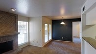 This single-story space showcases an open-concept living and dining area that feels bright and airy, thanks to multiple windows fitted with horizontal blinds and a set of glass-paned French doors that invite natural light throughout the day. The living area features wall-to-wall carpet in a neutral tan tone, providing a warm and comfortable foundation underfoot.

A standout architectural detail is the bold, deep navy accent wall in the dining area, which creates a striking contrast against the surrounding neutral white walls. A classic pendant light hangs above this space, adding a focal point and functional overhead lighting to the dining zone. The textured popcorn ceiling adds a layer of visual depth while maintaining a traditional aesthetic throughout.

The open layout allows the living and dining areas to flow seamlessly into one another, with a doorway visible leading to additional rooms beyond. The combination of ample natural light, neutral tones, and the dramatic accent wall creates a balanced ambiance that is both functional and visually interesting, making this a versatile and welcoming living space.