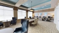 This cyber café offers a thoughtfully designed space that seamlessly blends comfort and functionality. The room features a distinctive tray ceiling with a striking teal accent, recessed lighting, and elegant pendant lights that create a warm, inviting ambiance throughout.

At the center of the space sits a large communal table surrounded by a mix of upholstered patterned chairs, providing a versatile area for collaboration or individual use. Complementing this arrangement are two high-back leather accent chairs, adding a sophisticated lounge element to the room.

Along the back wall, a built-in workstation counter houses multiple computer monitors, offering dedicated terminals for connected productivity. Additional pendant lighting above this area ensures a well-lit workspace. Framed artwork adorns the walls, enhancing the polished aesthetic of the room.

Large windows dressed with patterned valances and white blinds allow ample natural light to fill the space, creating a bright and airy atmosphere. Neutral-toned carpeting ties the room together, while the warm wall colors and teal ceiling details contribute to an upscale yet welcoming environment. This café is well-suited for both focused work and casual interaction.